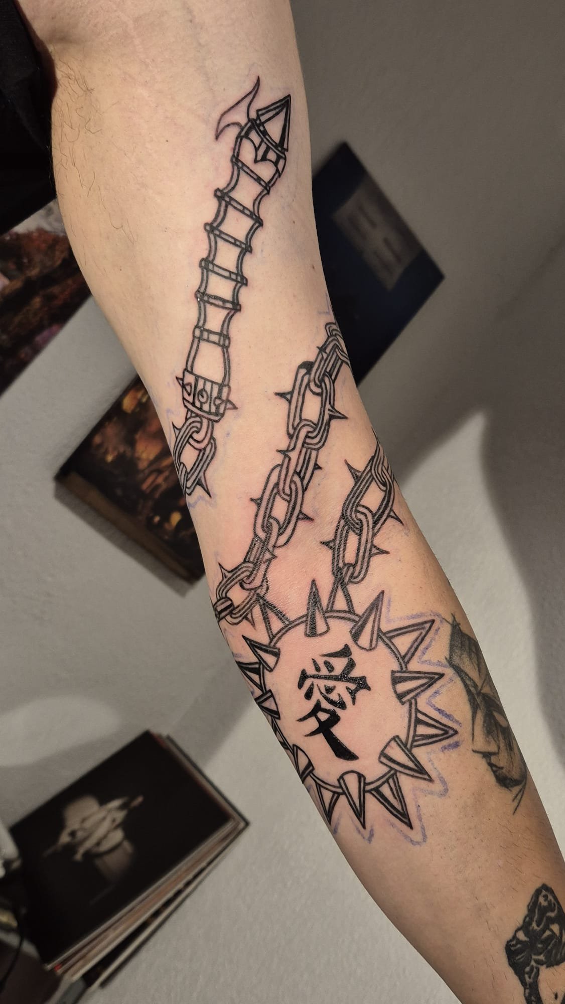 Antebraço com corrente de espigões a descer até um sol com o kanji de amor, tudo em blackwork